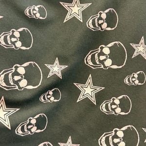 May include: Dark green fabric featuring a repeating pattern of white skulls and star shapes. The skulls are detailed with eye sockets and teeth, and the stars have a five-point design. The overall aesthetic is edgy and bold.
