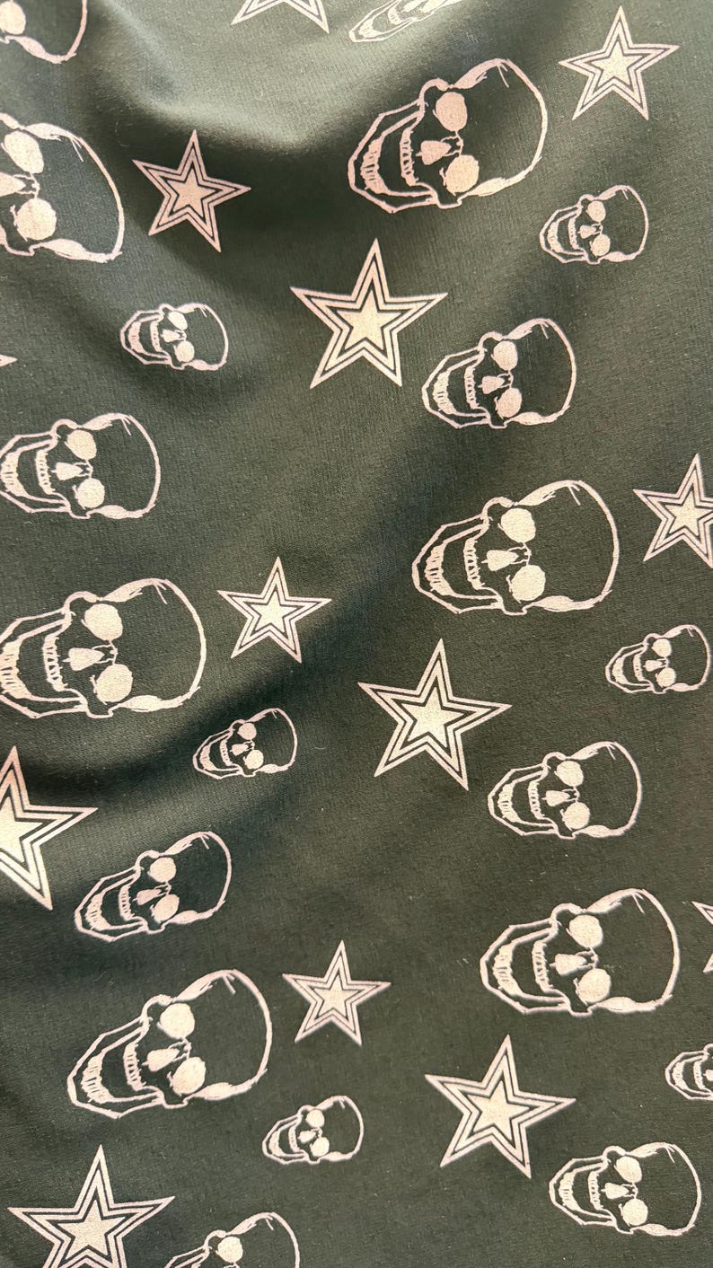 May include: Dark green fabric featuring a repeating pattern of white skulls and star shapes. The skulls are detailed with eye sockets and teeth, and the stars have a five-point design. The overall aesthetic is edgy and bold.