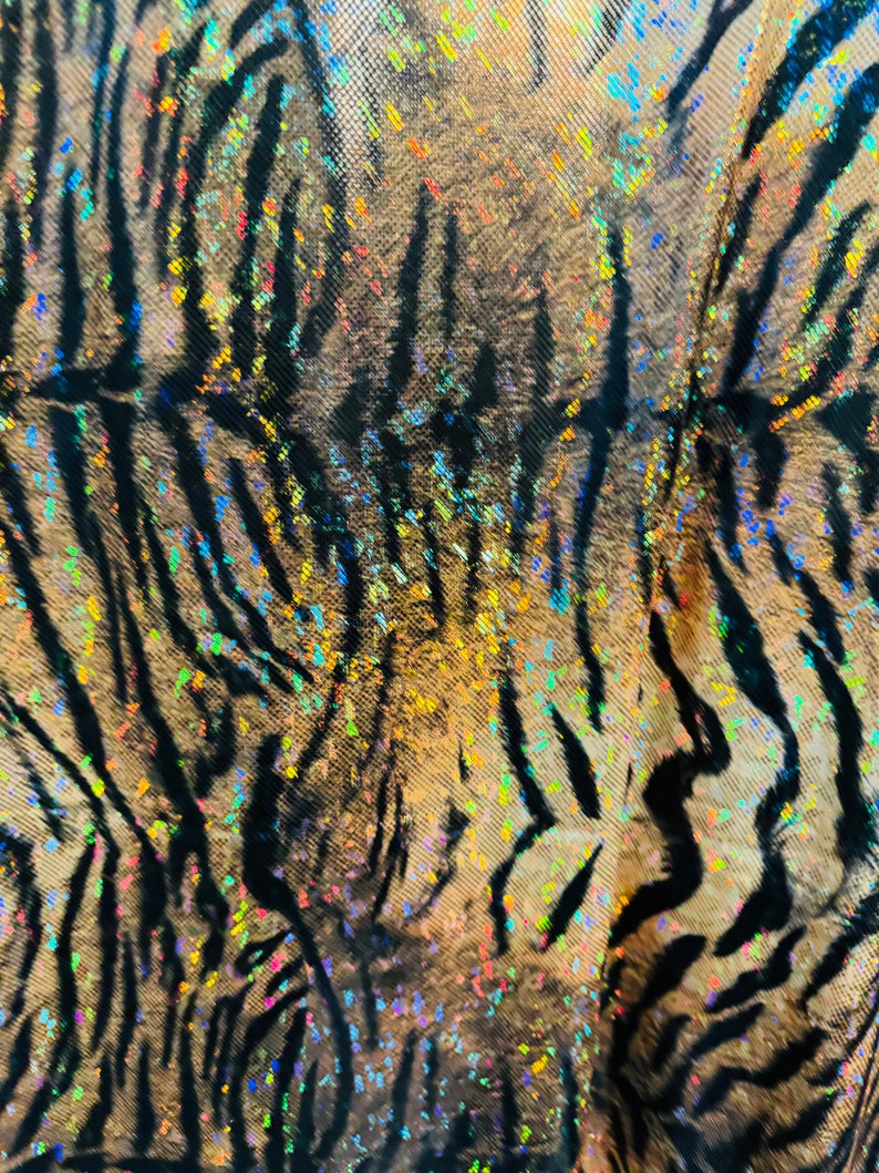 May include: Close-up of a fabric with a tiger stripe pattern. The fabric is iridescent and has a glittery finish. The stripes are black and the background is a light brown color.