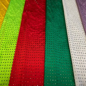 Op de afbeelding: Een verzameling verticale stroken stof in diverse kleuren, waaronder geel, limoengroen, rood, donkergroen, wit, lavendel en blauw. Elke strook is voorzien van een uniform rasterpatroon van kleine, reflecterende, ronde studs op het oppervlak.