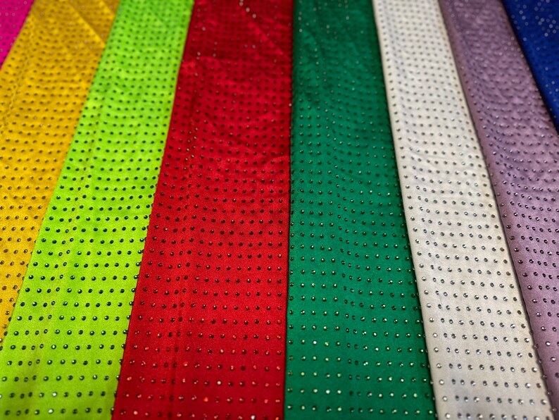 Op de afbeelding: Een verzameling verticale stroken stof in diverse kleuren, waaronder geel, limoengroen, rood, donkergroen, wit, lavendel en blauw. Elke strook is voorzien van een uniform rasterpatroon van kleine, reflecterende, ronde studs op het oppervlak.