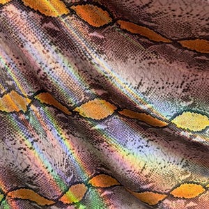 Puede incluir: Una tela con estampado de piel de serpiente y acabado holográfico. El material presenta una textura repetitiva en forma de diamante en tonos naranja, amarillo y morado, con un brillo reflectante que crea un efecto de luz de arcoíris.
