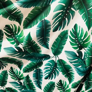 Op de afbeelding: Witte stof met een herhalend patroon van groene tropische bladeren. De bladeren zijn in verschillende maten en vormen, waaronder grote monstera bladeren en kleinere palmbladeren.