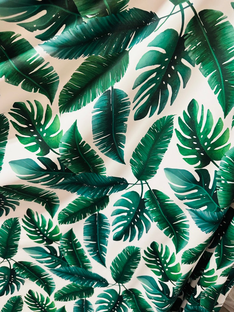 Op de afbeelding: Witte stof met een herhalend patroon van groene tropische bladeren. De bladeren zijn in verschillende maten en vormen, waaronder grote monstera bladeren en kleinere palmbladeren.