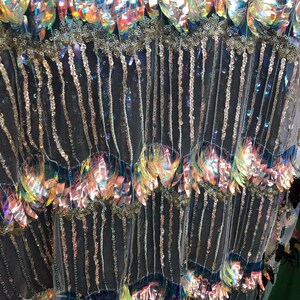 K&ouml;nnte beinhalten: Nahaufnahme eines irisierenden Stoffes mit Pailletten und Perlen. Der Stoff hat eine schwarze Netzbasis mit Reihen von irisierenden Pailletten und Perlen. Die Pailletten sind in einem Muster &uuml;berlappender Bl&auml;tter angeordnet.