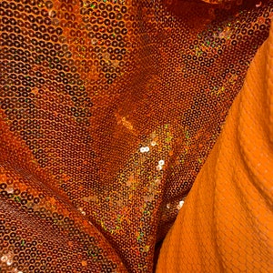 May include: Close-up of a fabric with a textured, iridescent orange sequin pattern. The fabric is folded over, revealing a smooth, solid orange fabric underneath.