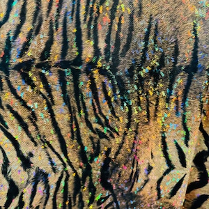 May include: Close-up of a fabric with a tiger stripe pattern. The fabric is iridescent and has a glittery finish. The stripes are black and the background is a light brown color.