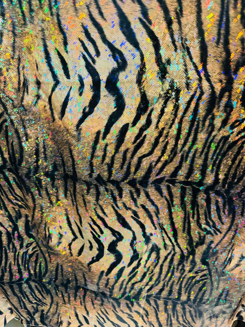May include: A close-up of a fabric with a tiger stripe pattern. The fabric is a light brown color with black stripes and iridescent glitter accents.