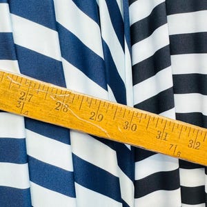 K&ouml;nnte beinhalten: Nahaufnahme eines Stoffes mit kr&auml;ftigen, vertikalen Streifen in Marineblau, Wei&szlig; und Schwarz. Ein Holzlineal liegt diagonal &uuml;ber dem Stoff und zeigt Messungen in Zentimetern an.