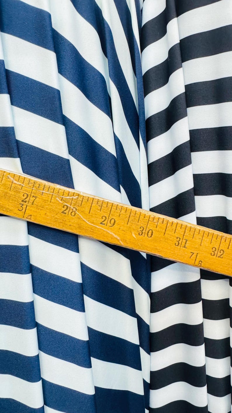 K&ouml;nnte beinhalten: Nahaufnahme eines Stoffes mit kr&auml;ftigen, vertikalen Streifen in Marineblau, Wei&szlig; und Schwarz. Ein Holzlineal liegt diagonal &uuml;ber dem Stoff und zeigt Messungen in Zentimetern an.