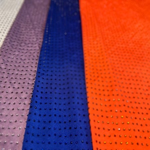 Op de afbeelding: Vier verticale stroken stof in groen, wit, paars, blauw en oranje, elk voorzien van een dicht, uniform patroon van kleine, donkere, verhoogde studs of strass-steentjes die het gehele oppervlak van het materiaal in een raster bedekken.