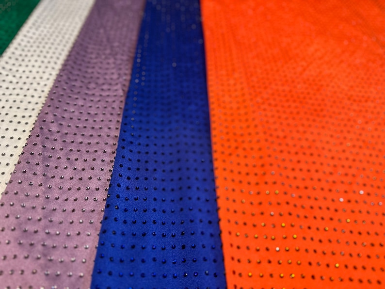 Op de afbeelding: Vier verticale stroken stof in groen, wit, paars, blauw en oranje, elk voorzien van een dicht, uniform patroon van kleine, donkere, verhoogde studs of strass-steentjes die het gehele oppervlak van het materiaal in een raster bedekken.