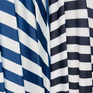 K&ouml;nnte beinhalten: Nahaufnahme von zwei Stoffen mit kr&auml;ftigen, diagonalen Streifen. Ein Stoff hat marineblaue und wei&szlig;e Streifen, w&auml;hrend der andere schwarz-wei&szlig;e Streifen aufweist. Die Stoffe sind drapiert und erzeugen weiche Falten und Schatten.