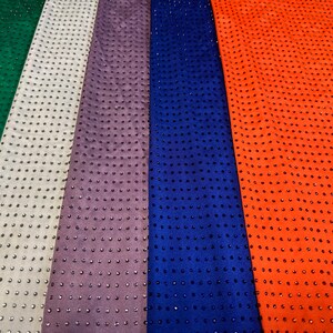 Op de afbeelding: Een collectie stoffen stroken in rood, groen, wit, paars, blauw en oranje, elk voorzien van een uniform raster van kleine, donkere, reflecterende studs die over het gehele oppervlak van het materiaal zijn ingebed, wat een textuur en patroon cre&euml;ert.