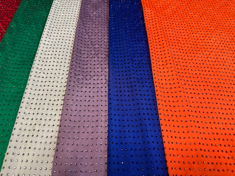 Op de afbeelding: Een collectie stoffen stroken in rood, groen, wit, paars, blauw en oranje, elk voorzien van een uniform raster van kleine, donkere, reflecterende studs die over het gehele oppervlak van het materiaal zijn ingebed, wat een textuur en patroon cre&euml;ert.