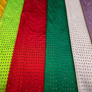 Op de afbeelding: Een collectie stoffen stroken naast elkaar in geel, limoengroen, rood, donkergroen, wit, lavendel en blauw. Elke strook is voorzien van een uniform raster van kleine, reflecterende, ronde studs die over het gehele oppervlak zijn aangebracht.