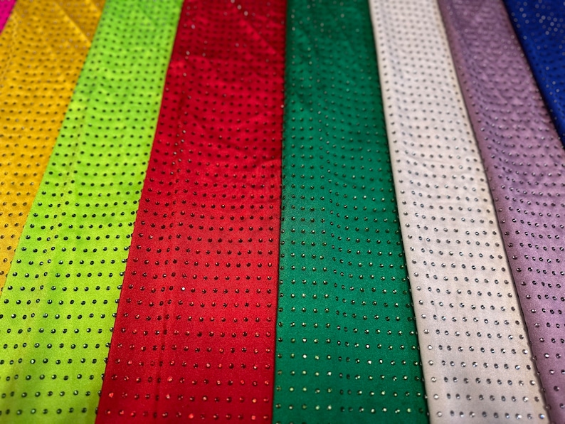 Op de afbeelding: Een collectie stoffen stroken naast elkaar in geel, limoengroen, rood, donkergroen, wit, lavendel en blauw. Elke strook is voorzien van een uniform raster van kleine, reflecterende, ronde studs die over het gehele oppervlak zijn aangebracht.