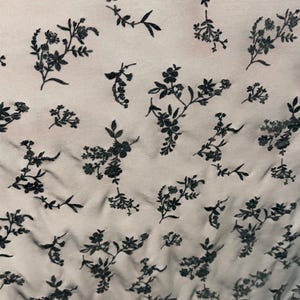 以下が含まれることがあります： 花柄のシアー生地のクローズアップ。 デザインは、明るいベージュの背景に散らばる繊細な黒い花と葉を特徴としています。 生地は柔らかく流れるような外観で、衣類や装飾目的で使用できる可能性があります。