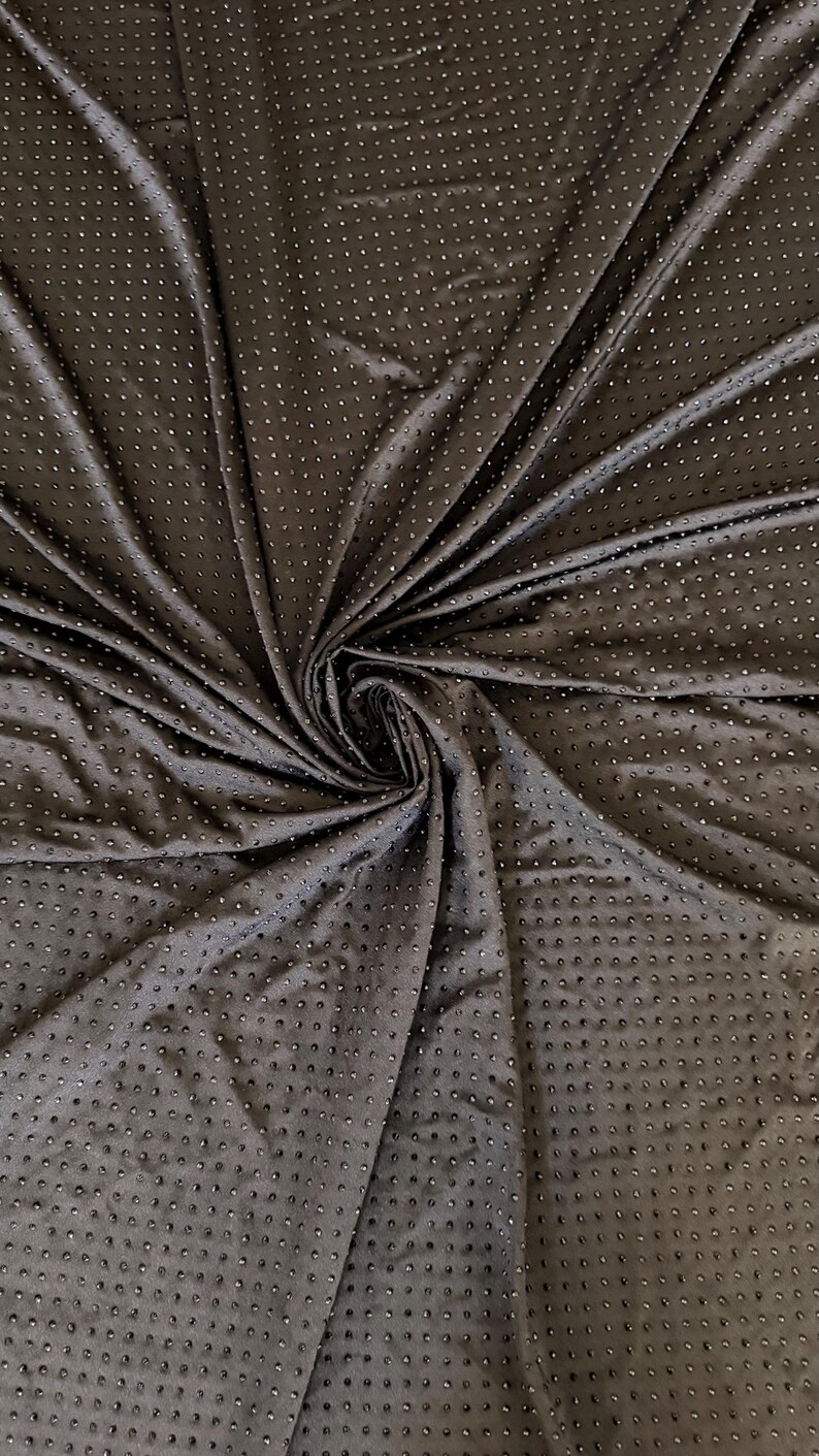 Op de afbeelding: Een donkerbruine stof met een dicht, uniform patroon van kleine, reflecterende, ronde studs gerangschikt in een raster. Het materiaal is in het midden samengebracht, waardoor stralende plooien en draperingen ontstaan die de textuur benadrukken.