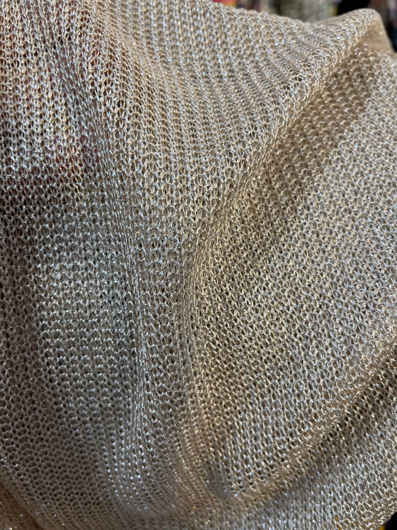 Puede incluir: Primer plano de una tela de punto beige con un acabado brillante. La tela tiene un tejido suelto y abierto y una superficie ligeramente texturizada.
