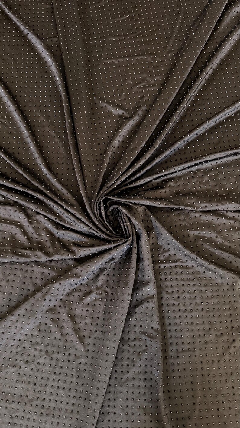 Op de afbeelding: Een donkerbruine stof met een dicht, uniform patroon van kleine, verhoogde, cirkelvormige stippen over het gehele oppervlak. Het materiaal is in een centrale werveling verzameld, waardoor diepe plooien en schaduwen ontstaan.