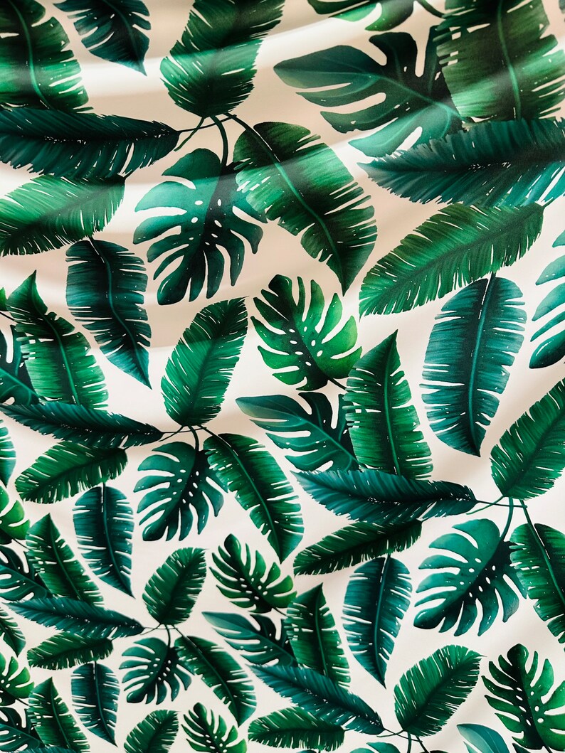 Op de afbeelding: Een witte stof met een herhalend patroon van groene tropische bladeren. De bladeren zijn in verschillende vormen en maten, waaronder palmbladeren en monstera bladeren.