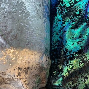 Op de afbeelding: Twee stofstalen, een zilverkleurig met een textuur en de andere iriserend groen en blauw met een textuur.