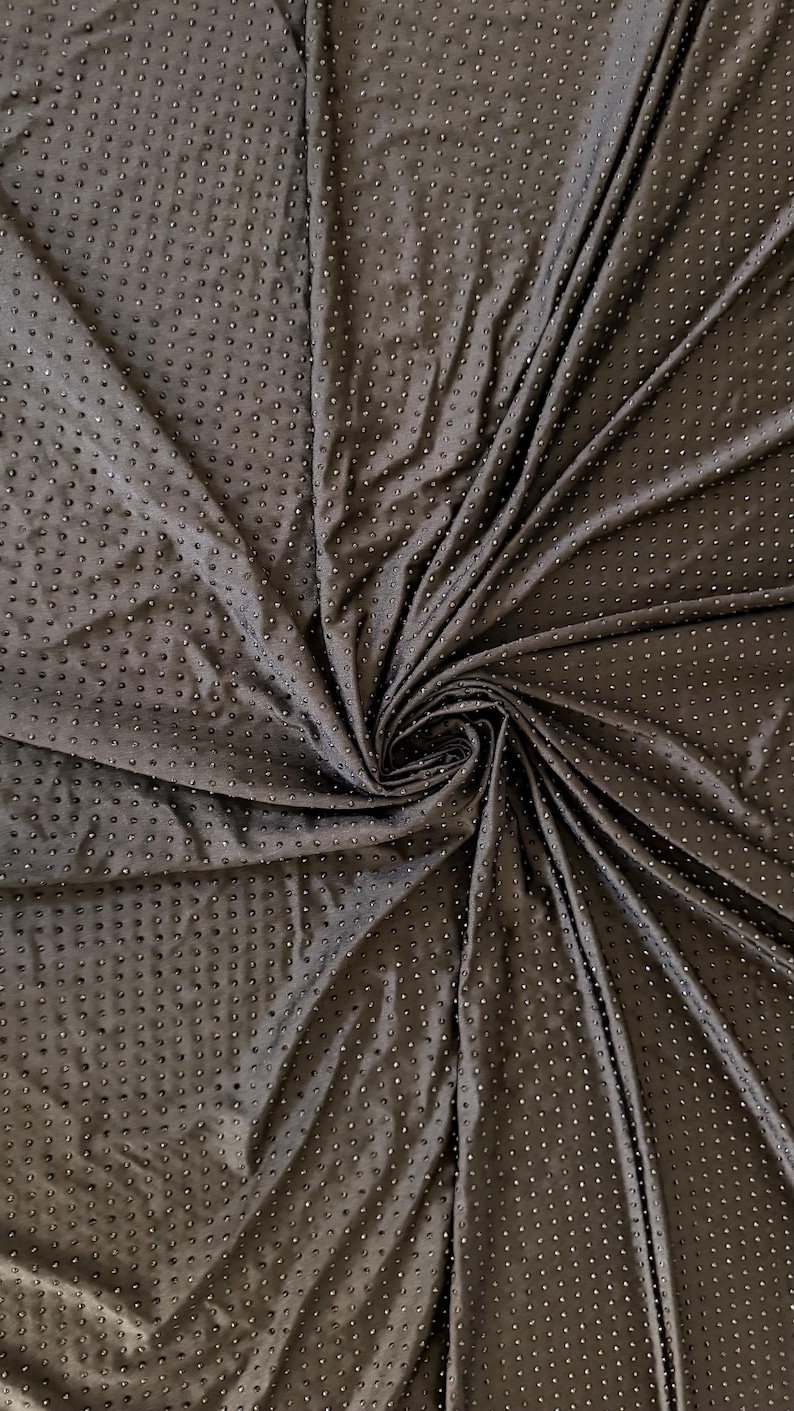 Op de afbeelding: Een donkerbruine stof met een herhalend patroon van kleine, verhoogde, cirkelvormige stippen in een raster. Het materiaal is in een centrale spiraal gedrapeerd, wat diepe plooien en schaduwen cre&euml;ert die de textuur van het oppervlak benadrukken.