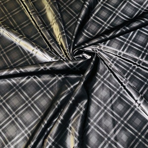 May include: A dark gray fabric with a plaid pattern. The fabric has a shiny, satin-like finish, with a repeating diamond pattern in shades of gray and white. The fabric is draped and folded, creating shadows and highlights.