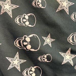 May include: Dark fabric featuring a repeating pattern of white skulls and five-pointed stars. The skulls have eye sockets, a nose cavity, and teeth. The stars have a double outline.