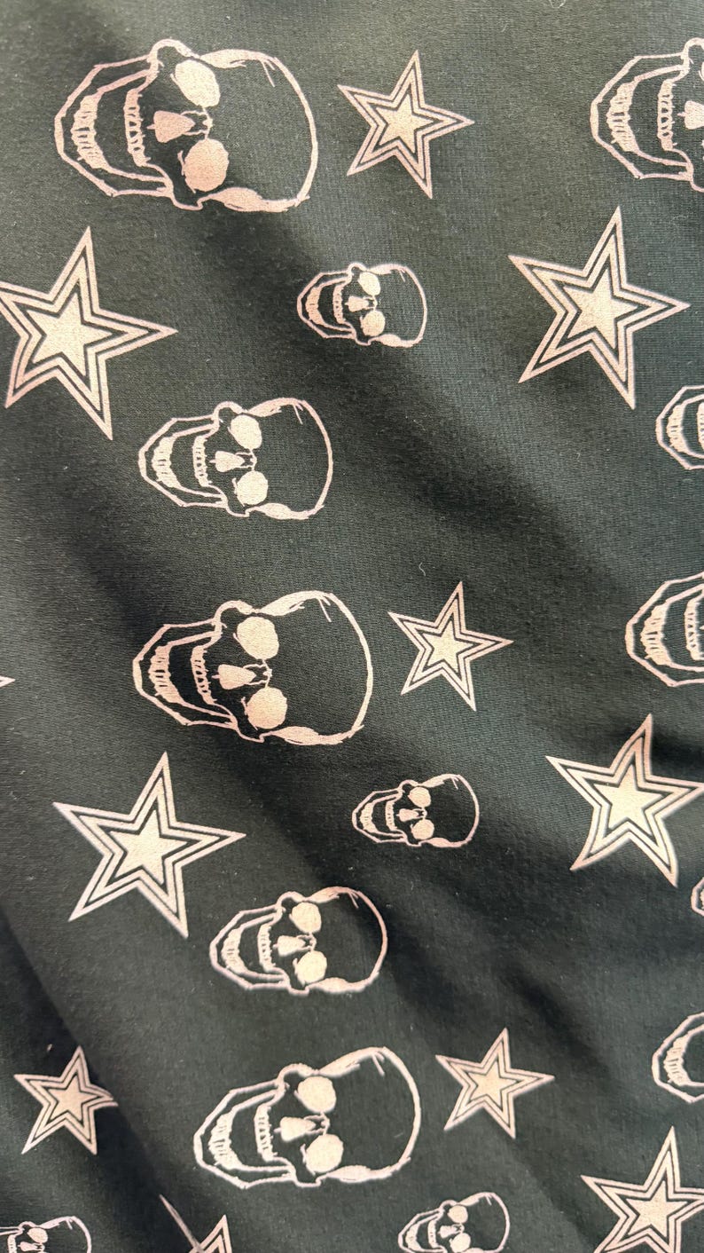 May include: Dark fabric featuring a repeating pattern of white skulls and five-pointed stars. The skulls have eye sockets, a nose cavity, and teeth. The stars have a double outline.