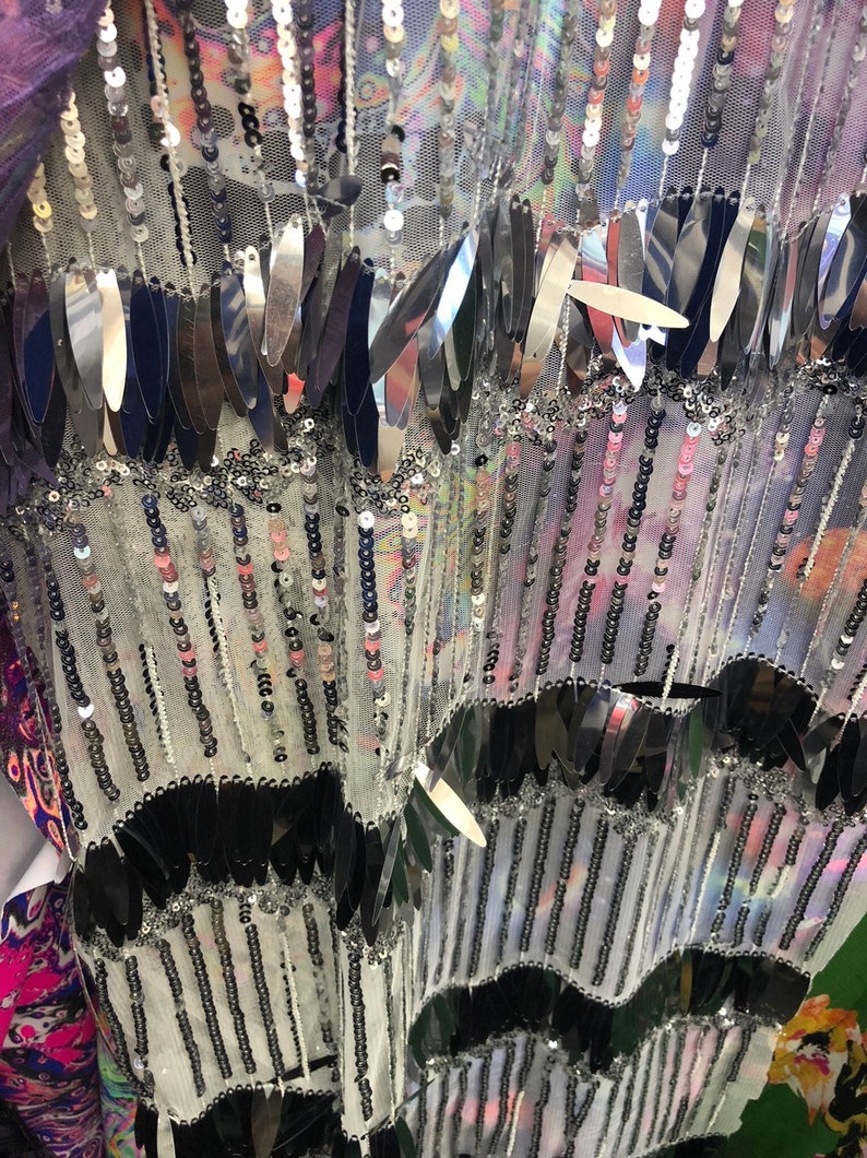 K&ouml;nnte beinhalten: Nahaufnahme eines silbernen und schwarzen Paillettenstoffes mit einem mehrschichtigen Design. Der Stoff hat eine durchsichtige, netzartige Textur und zeigt Reihen von silbernen Pailletten und schwarzen Fransen.