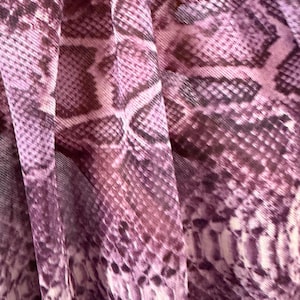 Könnte beinhalten: Ein leichter Stoff mit einem sich wiederholenden Schlangenhautmuster in verschiedenen Violett-, Mauve- und dunklen Pflaumentönen. Das Material ist in weichen, vertikalen Falten gerafft, was ein strukturiertes Aussehen mit Tiefe erzeugt.