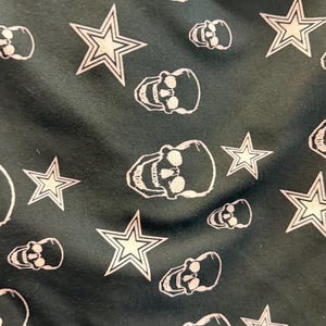 May include: Dark green fabric featuring a repeating pattern of white skulls and five-pointed stars. The skulls have eye sockets, a nose cavity, and teeth. The stars have a white outline and a filled-in centre.