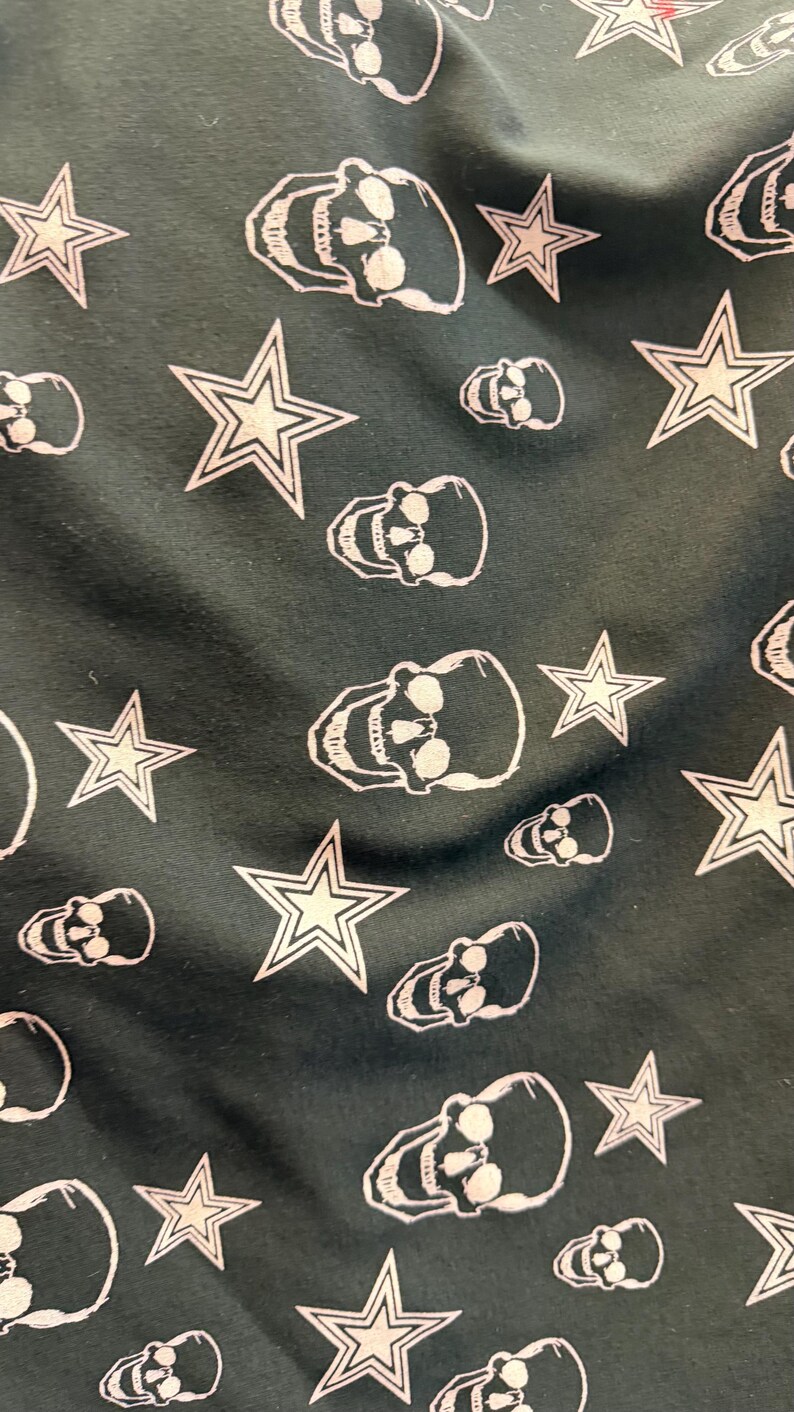 May include: Dark green fabric featuring a repeating pattern of white skulls and five-pointed stars. The skulls have eye sockets, a nose cavity, and teeth. The stars have a white outline and a filled-in centre.