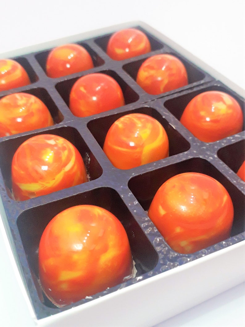 Puede incluir: Una caja de 10 chocolates de color naranja y amarillo en espiral dispuestos en forma de cuadr&iacute;cula. Los chocolates est&aacute;n en una caja blanca con divisores negros.