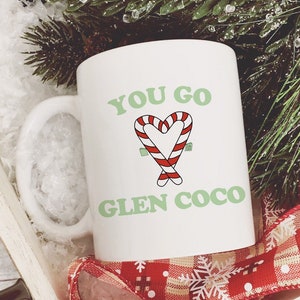 以下が含まれることがあります： 白地に緑と赤のキャンディーケーンのハートのデザインと「You go Glen Coco」の文字が書かれた白いセラミックマグカップ。マグカップは、赤と白のチェック柄のリボンがついた白い木製のトレイに入っています。