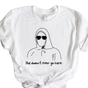 May include: White t-shirt with a black line drawing of a person wearing a hooded sweatshirt and sunglasses. The text "She doesn't even go here." is printed below the drawing.