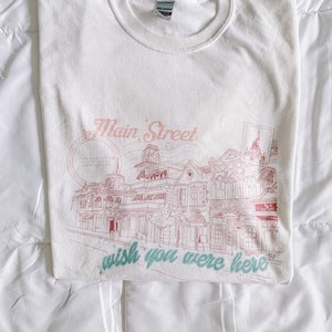 May include: White t-shirt with a pink graphic of Main Street, U.S.A. at Walt Disney World. The graphic includes the text "wish you were here".