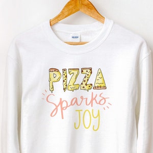 May include: White sweatshirt with a graphic design of the word "PIZZA" made of pizza slices. The text "Sparks Joy" is written below the word "PIZZA" in pink and yellow.