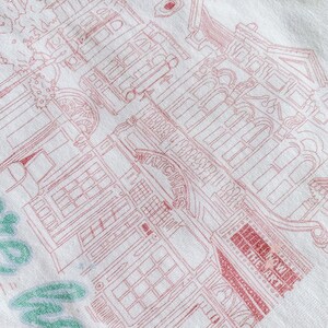 May include: A white sweatshirt with a pink outline of a building with the text "London Underground" and "The Market" on it. The text "Hello" is written in green on the bottom left corner of the sweatshirt.