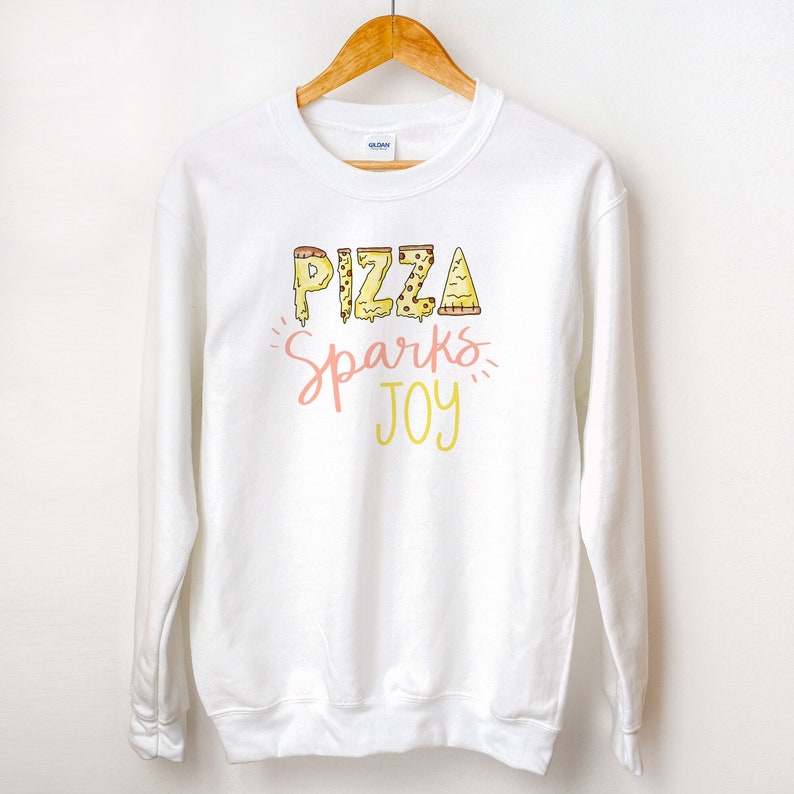 May include: White sweatshirt with a graphic design of the word "PIZZA" made of pizza slices. The text "Sparks Joy" is written below the word "PIZZA" in pink and yellow.