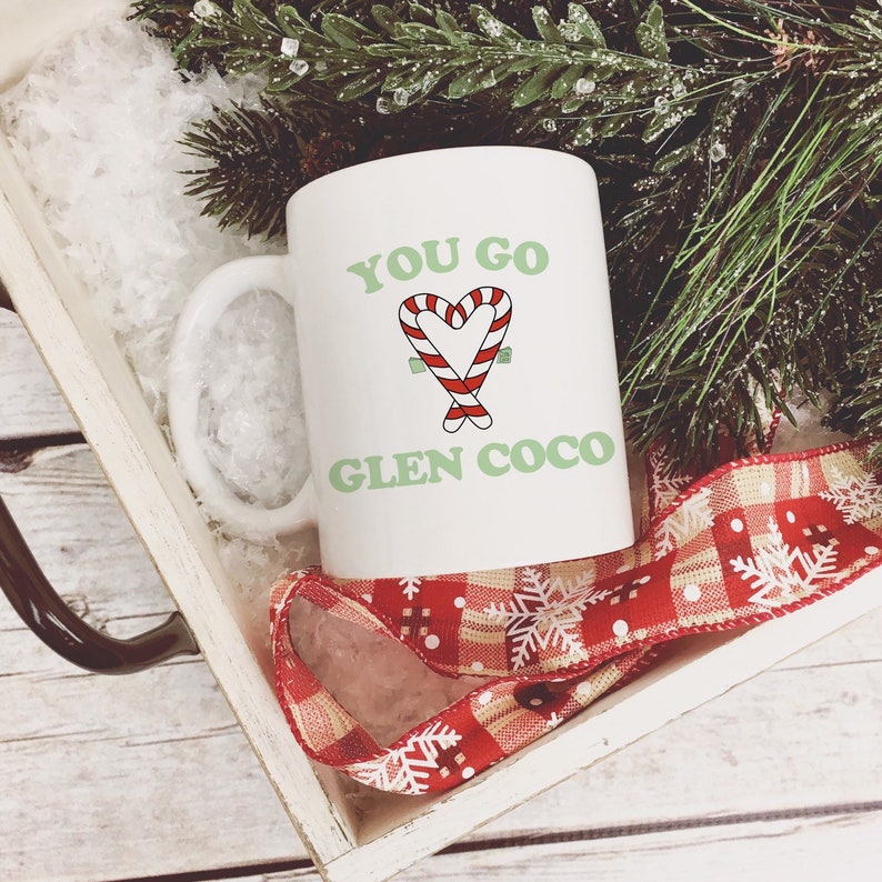 以下が含まれることがあります： 白地に緑と赤のキャンディーケーンのハートのデザインと「You go Glen Coco」の文字が書かれた白いセラミックマグカップ。マグカップは、赤と白のチェック柄のリボンがついた白い木製のトレイに入っています。