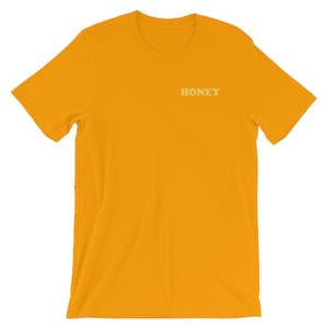 Pode incluir: Uma camiseta amarela de manga curta com a palavra "HONEY" impressa em amarelo claro na frente.