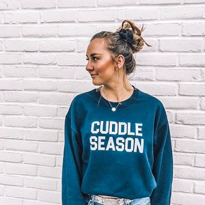 May include: A woman wearing a navy blue sweatshirt with the text "Cuddle Season" in white lettering. She is wearing blue jeans and a silver belt.