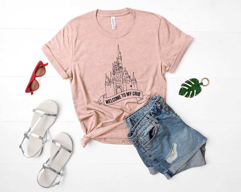 May include: A pink t-shirt with a black outline of Cinderella's castle and the text "Welcome to my crib".