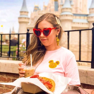 May include: A person wearing red heart-shaped sunglasses and a light pink t-shirt with a yellow Mickey Mouse waffle design. The person is holding a drink with a straw and a waffle with strawberries.