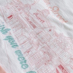 May include: A white t-shirt with a pink and green design. The design features a building with the text "Main Street" and "We were here" in a cursive font.