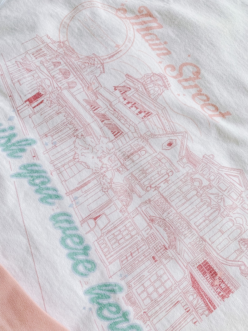 May include: A white t-shirt with a pink and green design. The design features a building with the text "Main Street" and "We were here" in a cursive font.