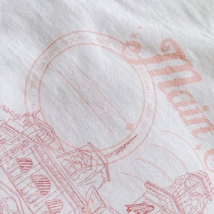May include: A white t-shirt with a pink graphic design of a building with a circle stamp that says "Established 1982" and the text "Atlantic City" in a cursive font.